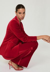 Rød fløjlssuit med tætsiddende jakke og matchende bukser, der har knapdetaljer og kontrasterende sidelameller. Båret med røde strophæle.