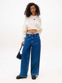 White knitted cropped sweater with ribbed texture, paired with high-waisted wide-leg blue denim jeans. Model holds a small black bag.