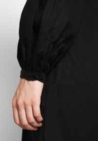 Schwarzes Kleid aus strukturiertem Stoff, mit puffärmel und einem Knopfmanschetten-Detail, sowie einer Nahaufnahme der Hand, die auf dem Kleid ruht.