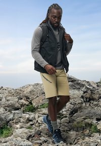 Must have sansleeve vest musta hallita halli pikka käistega topp, beežid lühikesed püksid ja hallid jalatsid. Seismine kivisel maastikul sinise taeva all.