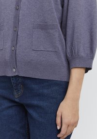 Lilla kudumine kardigan kahe ees oleva taskuga, nööbivarrukate ja soonikkangaga, koos tumedast sinisest denimist teksadega.