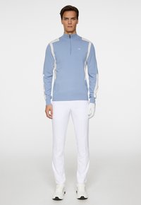 Ljusblå zip-up tröja med vita detaljer, långa ärmar, ribbade muddar, kombinerad med vita byxor och sneakers. Enkelt logotyp på bröstet.