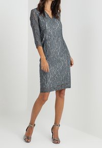 Robe en dentelle grise avec des manches trois-quarts et un décolleté en V, présentant un motif floral et un ourlet droit. Assortie avec des talons à brides de cheville argentés.