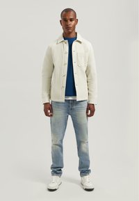 Giacca button-up bianca texturizzata sopra un maglione blu a maglia, abbinata a jeans blu chiaro e sneakers bianche. In piedi davanti a uno sfondo neutro.
