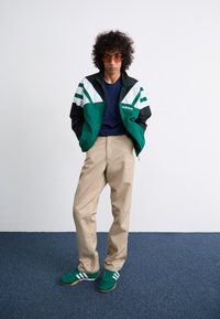 Casaco corta-vento verde e preto com riscas brancas, camisa azul-marinho, calças bege e sapatos verdes com detalhes brancos; usado com óculos de sol.