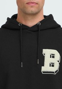 Uomo che indossa una felpa nera con cappuccio, con una lettera "B" testurizzata color panna sul petto e coulisse con punte in metallo.