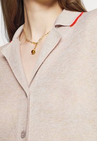 Gros plan d'une personne portant une chemise beige boutonnée avec un bord rouge au col et un collier en chaîne dorée avec un pendentif en ambre.