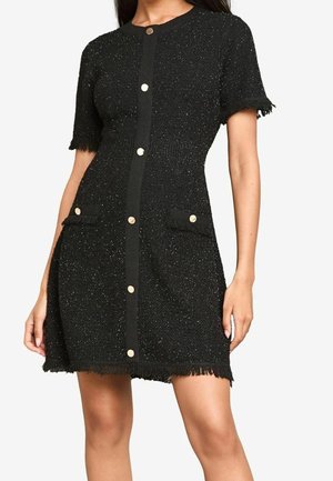 Musta lyhythihainen neulemekko, jossa on etuosassa kultaiset napit, kaksi etutaskua sekä hapsureunukset hihoissa, taskuissa ja helmassa.