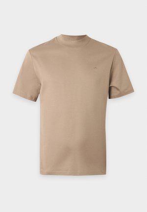 Homme debout portant un t-shirt beige à manches courtes, un jean ample gris foncé et des chaussures noires, une main dans la poche.