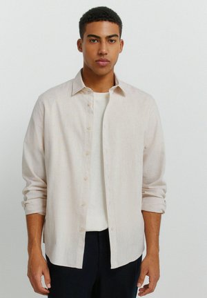 Młody mężczyzna o krótkich kręconych włosach, ubrany w jasnobeżową koszulę zapinaną na guziki, na białej koszulce i ciemnych spodniach, stoi na jednolitym tle.