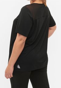 Donna che indossa un top nero a maniche corte e largo con un yoke trasparente sulla schiena e pantaloni neri, in piedi con una mano su un fianco.