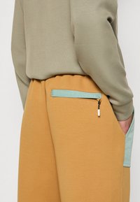 Beige Jogginghose mit einer mintgrünen Tasche, Reißverschluss und strukturiertem Stoff, kombiniert mit einem hellgrünen Sweatshirt.