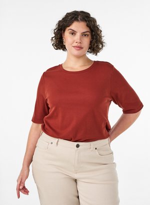 Donna con capelli ricci che indossa una camicia a maniche corte color ruggine e pantaloni beige chiaro, in piedi con una mano sui fianchi davanti a uno sfondo bianco.