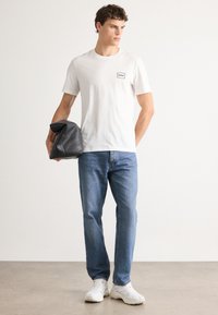 Ung mann i hvit Hugo T-skjorte, blå jeans, hvite sneakers, holder en brettet svart lærjakke, står på et lyst gulv mot en hvit vegg.