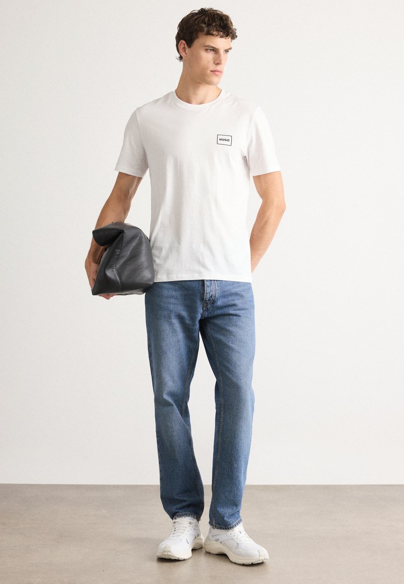 Ung mand iført hvid Hugo T-shirt, blå jeans, hvide sneakers, holder en foldet sort læderjakke, står på et lyst gulv mod en hvid væg.