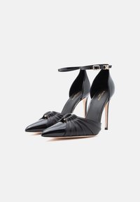 Mustad mustakontsadega stiletod, millel on terav lakktald, plekitud nahast ülaosa ja pahkluu rihmad kuldsete pandlatega, valgel taustal.