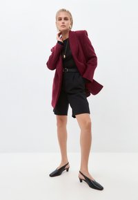 Blazer oversize bordeaux con una texture sottile, top nero aderente, pantaloni corti neri su misura e décolleté nere con tacco basso a blocco.