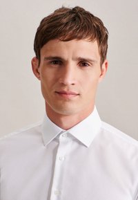 Witte gekraagde blouse met een gladde textuur en knoopsluiting aan de voorkant. Model heeft kort, lichtbruin haar en een neutrale uitdrukking.