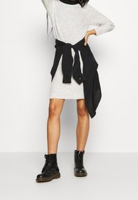 Grå stickad klänning med långa ärmar, svart skjorta knuten i midjan, och svarta snörkängor. Slät textur med kontrasterande färger.