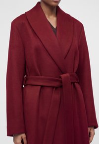 Femme portant un manteau en laine rouge profond ceinturé avec de larges revers, montrée du cou jusqu’à mi-cuisse sur un fond clair uni.