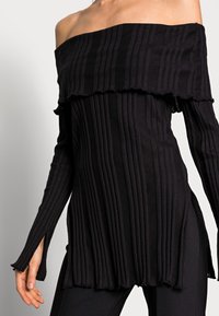 Černý žebrovaný pletený svetr s odhalenými rameny a dlouhými rukávy, které mají otvory na palce, s rozšířeným lemem, nošený s černými kalhotami.