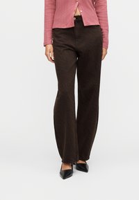 Femme portant un pantalon évasé à motifs brun foncé, un haut côtelé rose poussiéreux dévoilant le ventre, et des talons noirs à bouts pointus.