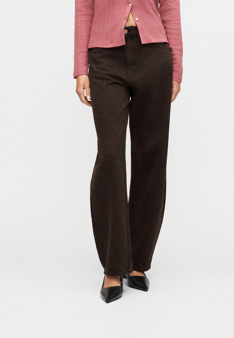 Femme portant un pantalon évasé à motifs brun foncé, un haut côtelé rose poussiéreux dévoilant le ventre, et des talons noirs à bouts pointus.