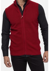Chaleco de punto rojo con cierre de cremallera, que presenta un patrón texturizado y un dobladillo acanalado. Lleva encima de una camiseta negra de manga larga, destacando su diseño.