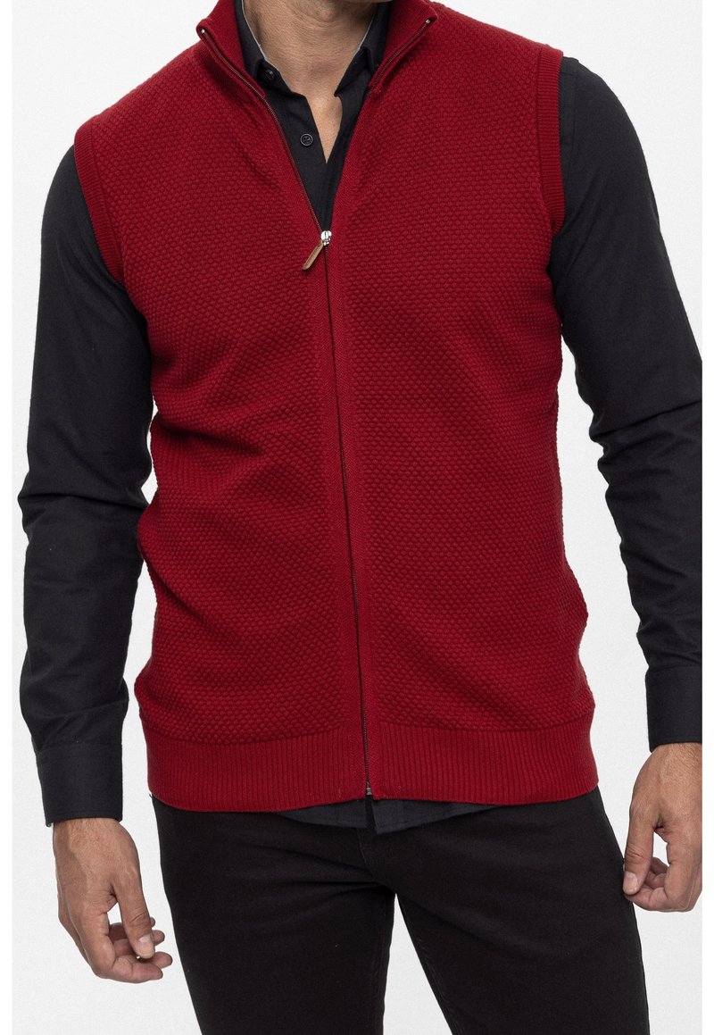 Chaleco de punto rojo con cierre de cremallera, que presenta un patrón texturizado y un dobladillo acanalado. Lleva encima de una camiseta negra de manga larga, destacando su diseño.