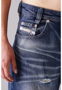 Blaue Jeans aus Denim mit einem ausgewaschenen Finish, abgetragenen Stellen, kupfernen Nieten und einem weißen Etikett auf der Gesäßtasche, das ein Logo zeigt.