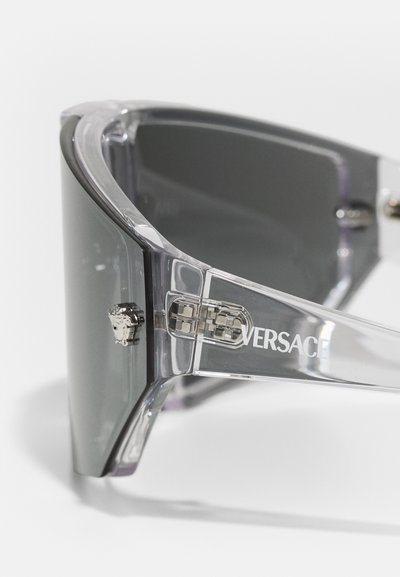 Transparent light gray sunglasses with bold angular shape, dark gradient lenses, and silver hardware featuring the Versace logo on the frame.