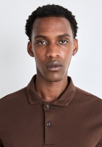 Brun polo-skjorte med klassisk krage, to-knapps lukkning, laget av glatt stoff. Har korte ermer og en skreddersydd passform.