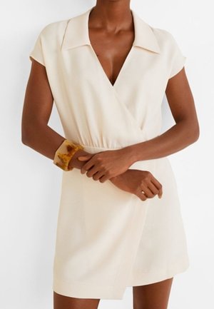 Femme portant une robe portefeuille à manches courtes couleur crème avec un col, accessoirisée d’un bracelet jonc en ambre marbré au poignet.