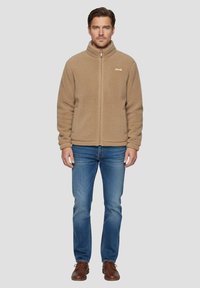 Giacca in pile color cammello con colletto alto, zip integrale e marchio tono su tono. Abbinata a jeans blu e scarpe in pelle marrone. Tessuto liscio.