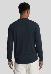 Uomo che indossa un maglione a maniche lunghe blu scuro e pantaloni beige chiaro, in piedi con la schiena rivolta verso lo spettatore, su uno sfondo neutro.