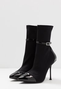 Botines negros de tela con una textura suave, que presentan punteras puntiagudas de charol y un delgado tacón de aguja, adornados con una hebilla.