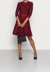 Röd klänning med svart grafisk text, trekvartslång ärm, åtdragen midja och utsvängt kjoldel. Matchas med en liten svart handväska och svarta klackar.