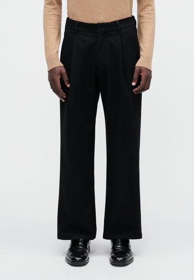 Pantalons noirs avec une coupe évasée, comportant des plis à l'avant et une texture lisse, associés à des chaussures noires et un haut beige à manches longues.