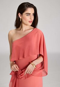 Ein abendliches Ein-Schulterkleid in Koralle, mit einem fließenden Chiffon-Overlay und gerafftem Stoffdetail an der Taille. Akzentuiert mit einem goldenen Armreif.