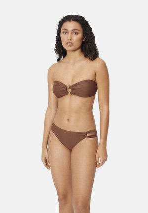 Mujer de pie mirando al frente, vestida con un bikini marrón sin tirantes, con aros dorados como detalles en la parte superior y recortes en los costados de la parte inferior.