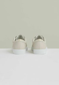 Sneakers bej cu un superior din piele texturată, talpă din cauciuc alb și branding discret pe călcâi. Design de profil jos cu formă rotundă.