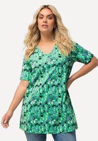 Vihreä V-pääntie tunika, jossa on rohkea geometrinen kuvio mustassa, purppurassa ja kermahiileessä. Lyhyet hihat ja rento istuvuus.