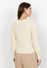 Cream knitted sweater with a ribbed texture, slightly cropped design, and long sleeves. Features a round neckline and subtle stitching details.