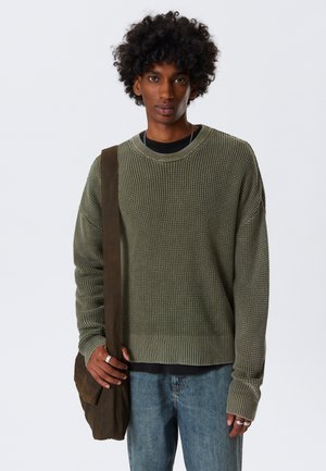 Jersey verde de punto con un patrón texturizado, combinado con una camiseta negra, jeans de color azul claro y un bolso marrón al hombro.