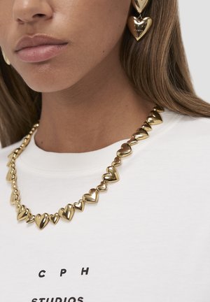 Close-up of a woman wearing gold heart-shaped earrings and matching necklace over a white shirt with "CPH STUDIOS" text.