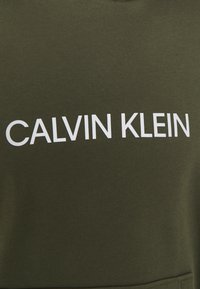 Olivengrøn bomulds-sweatshirt med fed hvid "CALVIN KLEIN" tekst tværs over brystet. Glat tekstur og klassisk rundhalset design.