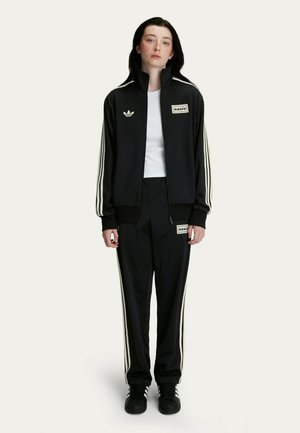 Black athletic tracksuit with white stripes, zip-up jacket, and pants featuring logo patches. Model wears a white shirt underneath.