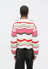 Άτομο με πλεξούδες φοράει πολύχρωμο πουλόβερ με κυματιστές ρίγες και μαύρο παντελόνι, στέκεται μπροστά σε απλό λευκό φόντο.