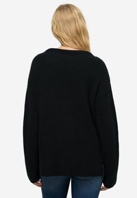 Schwarzer, oversized Strickpullover mit geripptem Muster, weitem runden Ausschnitt und abgesenkten Schultern, kombiniert mit blauen Jeans.