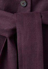 Tissu texturé violet foncé avec une ceinture attachée en nœud sous un bouton noir sur un vêtement.